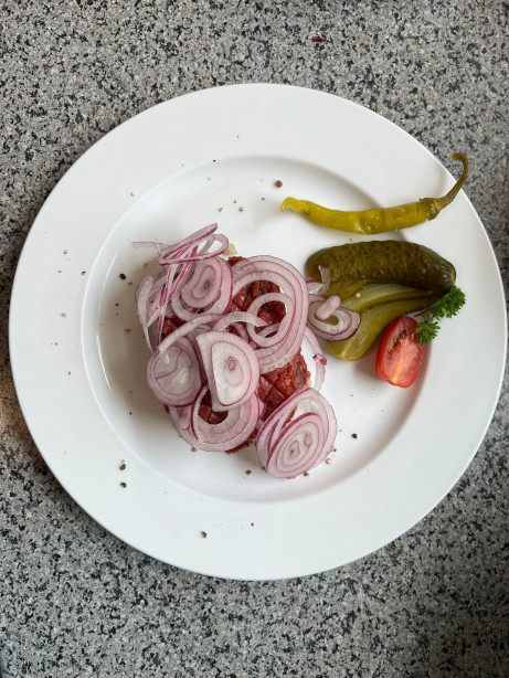 Das Bild zeigt einen typischen Hackepeter-Teller: linksseitig befindet sich ein Stück Brot mit Hackepeter darauf mit vielen rote Zwiebelscheiben garniert. Rechtsseitig am Tellerrand liegt eine grüne eingelegte Peperoni, eine längst aufgefächerte geschnittene saure Gurke und eine halbe Tomate mit Petersilie.